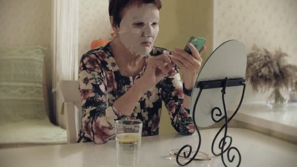 Uma mulher idosa olha em um espelho e aplica uma máscara de tecido cosmético em seu rosto. Ela usa o telefone. Cuidados faciais. Cirurgia plástica e injecções de colagénio. Imagens realistas com suas próprias imperfeições . — Vídeo de Stock
