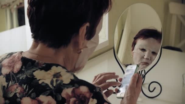 Una donna anziana si guarda allo specchio e applica una maschera di tessuto cosmetico sul viso. Usa il telefono. Cura del viso. Chirurgia plastica e iniezioni di collagene. Immagini realistiche con le proprie imperfezioni . — Video Stock