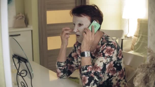 En gammal kvinna ser i en spegel och gäller en kosmetisk vävnad mask för ansiktet. Hon använder telefonen. Hudvård för ansiktet. Plastikkirurgi och kollagen injektioner. Realistiska bilder med sina egna brister. — Stockvideo