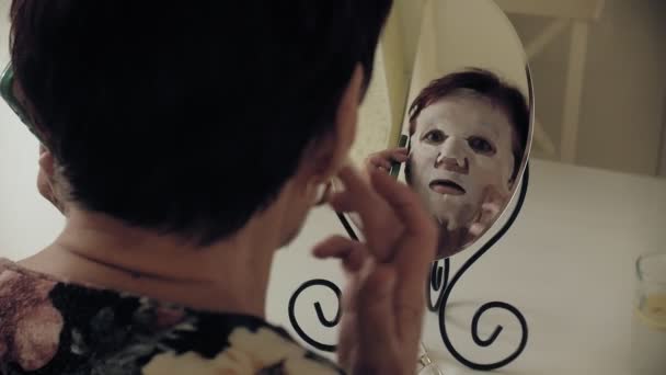Egy öregasszony néz a tükörbe, és a kozmetikai szövet maszk vonatkozik az arcát. Ő használ a telefon. Arcápoló. Plasztikai sebészet és kollagén injekciók. Reális képet a saját hiányosságai. — Stock videók