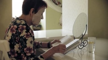 Yaşlı bir kadın masada oturur, kozmetik kumaş maskesi yüzüne karşı uygulanır. Bir kitap okuyor. Yüz bakımı. Plastik cerrahi ve Kollajen enjeksiyonları. Kendi kusurları ile gerçekçi görüntüler