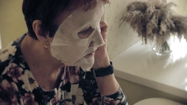 Μια ηλικιωμένη γυναίκα κοιτάζει στον καθρέφτη και εφαρμόζει μια μάσκα καλλυντικών ιστού στο πρόσωπό της. Χρησιμοποιεί το τηλέφωνο. Περιποίηση προσώπου. Πλαστική χειρουργική και ενέσεις κολλαγόνου. Ρεαλιστικές εικόνες με τις δικές τους ατέλειες. — Αρχείο Βίντεο