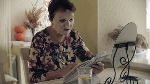 Een oude vrouw zit aan de tafel, een cosmetische stof masker is toegepast op haar gezicht. Ze leest een boek. Gelaatsverzorging. Plastische chirurgie en collageen injecties. Realistische beelden met hun eigen onvolkomenheden. — Stockvideo