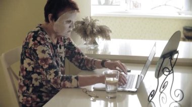 Yaşlı bir kadın masada oturur, kozmetik doku maskesi yüzüne karşı uygulanır. O bir bilgisayar kullanır. Yüz bakımı. Plastik cerrahi ve Kollajen enjeksiyonları. Kendi kusurları ile gerçekçi görüntüler.