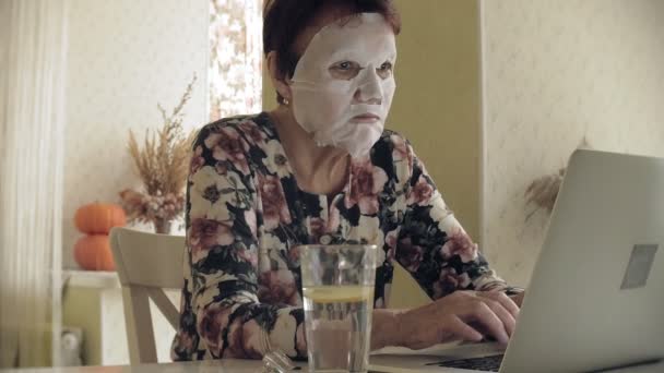 Een oude vrouw zit aan de tafel, een cosmetische weefsel masker is toegepast op haar gezicht. Ze maakt gebruik van een computer. Gelaatsverzorging. Plastische chirurgie en collageen injecties. Realistische beelden met hun eigen onvolkomenheden. — Stockvideo