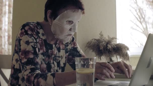 Stará žena sedí u stolu, kosmetické tkáně maska na tvář. Ona používá počítač. Péče o pleť. Plastická chirurgie a kolagenové injekce. Realistické obrazy s vlastní nedokonalosti. — Stock video