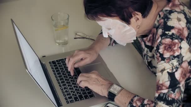 En gammal kvinna sitter vid bordet, en kosmetisk vävnad mask appliceras på hennes ansikte. Hon använder en dator. Hudvård för ansiktet. Plastikkirurgi och kollagen injektioner. Realistiska bilder med sina egna brister. — Stockvideo