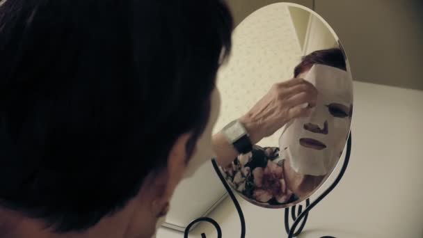 Egy idős nő úgy néz ki, egy tükör, kozmetikai szövet maszk eltávolítása az arcát. Arcápoló. Plasztikai sebészet és kollagén injekciók. Reális képet a saját hiányosságai. — Stock videók