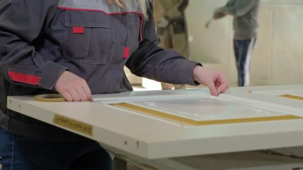 Una mujer en un taller pega una cinta protectora en la producción de puertas interiores de madera — Vídeos de Stock