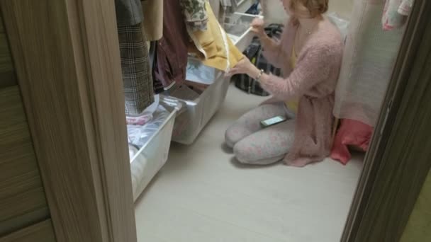 En vacker flicka leenden, sorterar och viker kläder i korgar i sitt omklädningsrum. sätter saker i ordning. använder smartphone — Stockvideo