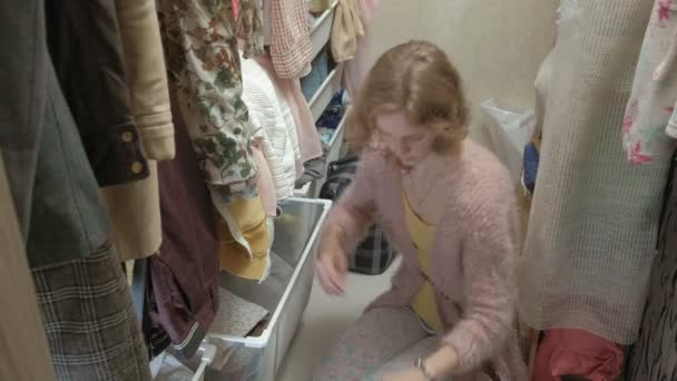 Een mooi meisje glimlacht, sorteert en plooien van kleren in manden in haar kleedkamer. Zet orde op zaken. maakt gebruik van de smartphone — Stockvideo