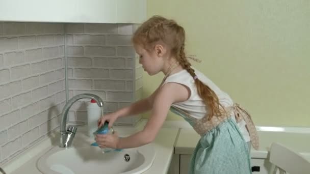 Beautiful little girl in an apron washes a mug in the bright kitchen, helps parents — Stock Video