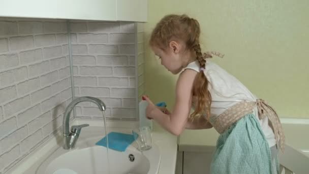 Menina bonita em um avental lava uma caneca na cozinha brilhante, ajuda os pais — Vídeo de Stock