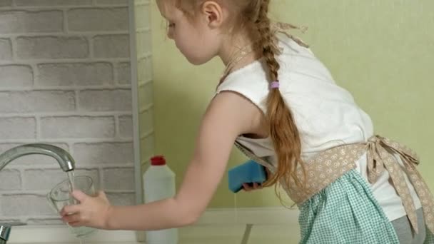 Menina bonita em um avental lava uma caneca na cozinha brilhante, ajuda os pais — Vídeo de Stock