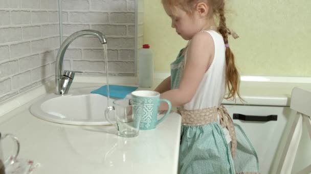 Όμορφο κοριτσάκι σε μια ποδιά στέκεται σε μια καρέκλα και πλένει μια κούπα στην φωτεινή κουζίνα, βοηθά τους γονείς — Αρχείο Βίντεο