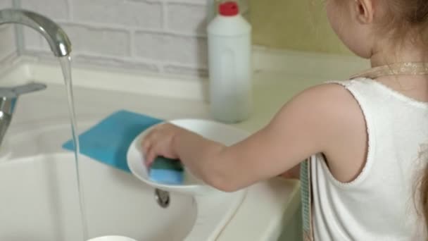 Belle petite fille dans un tablier est debout sur une chaise et lave une tasse dans la cuisine lumineuse, aide les parents — Video