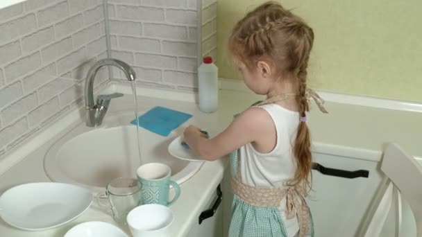 Красива маленька дівчинка в фартусі стоїть на стільці і миє посуд на світлій кухні, допомагаючи батькам — стокове відео