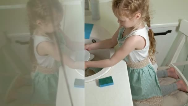 Belle petite fille dans un tablier est debout sur une chaise et laver la vaisselle dans la cuisine lumineuse, aider les parents — Video