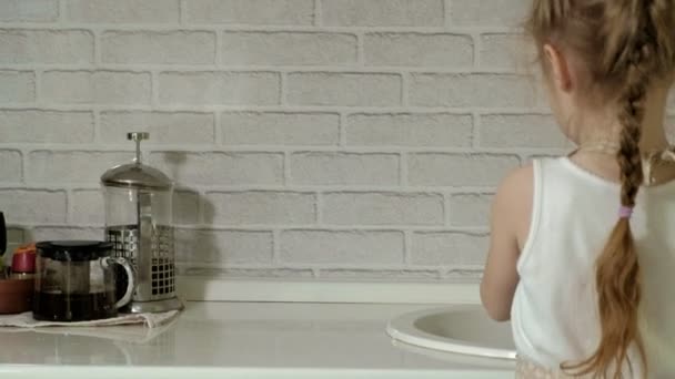 Vakker liten jente i et forkle står på en stol og tørker av bordet. Arbeidsflaten på det lyse kjøkkenet hjelper foreldrene dine. – stockvideo