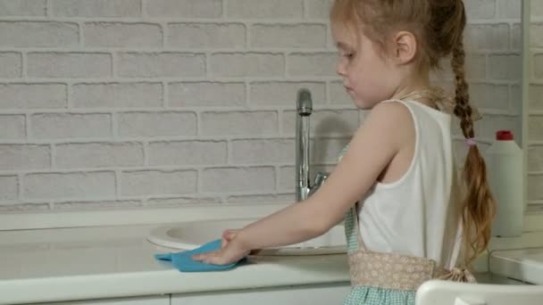 Όμορφο κοριτσάκι σε μια ποδιά στέκεται σε μια καρέκλα και σκουπίζει τον πίνακα, η επιφάνεια εργασίας στην κουζίνα φωτεινά βοηθά τους γονείς — Αρχείο Βίντεο