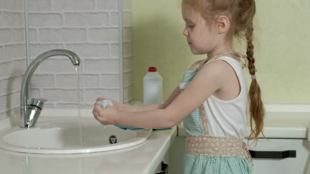 Beautiful little girl in an apron is standing on a chair in a bright kitchen, applies dish detergent to a sponge, helps parents — Stock Video