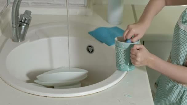 Menina bonita em um avental está de pé em uma cadeira, na cozinha brilhante, lava uma xícara com uma esponja, ajuda os pais — Vídeo de Stock