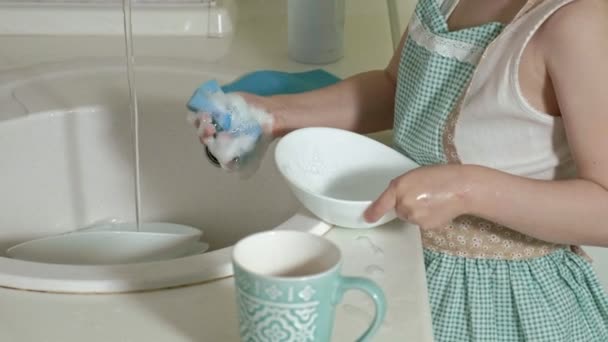 Belle petite fille dans un tablier est debout sur une chaise, dans la cuisine lumineuse, faire la vaisselle avec une éponge, aider les parents — Video