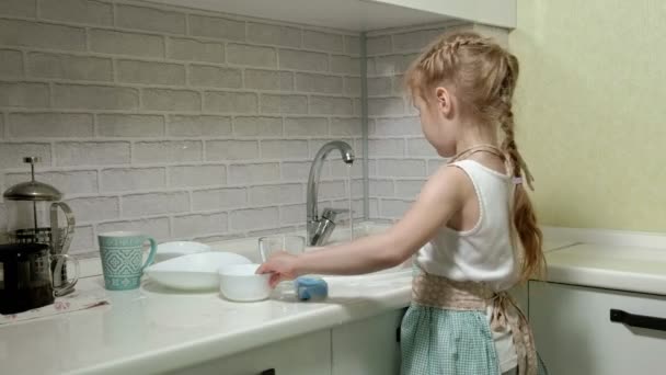 Красивая маленькая девочка в фартуке стоит на стуле, на яркой кухне, моет посуду губкой, помогает родителям — стоковое видео