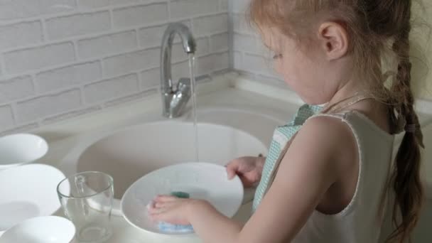 Mooi meisje in een schort is permanent op een stoel, in de lichte keuken, afwassen met een spons, ouders helpen — Stockvideo