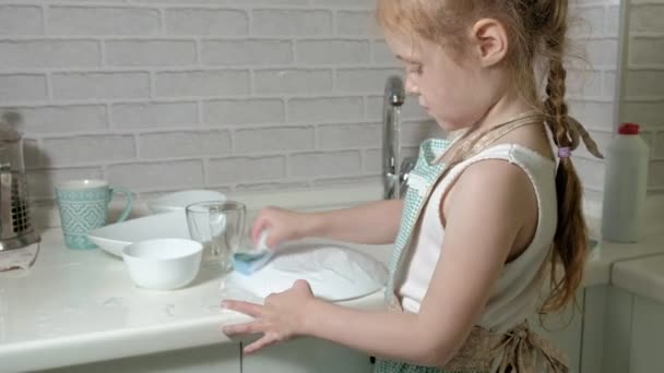 エプロンの中の美しい少女は両親の手伝いは、スポンジで皿を洗って明るいキッチンの椅子に立っています。 — ストック動画