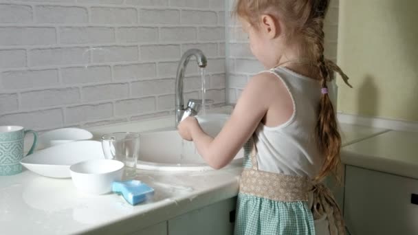 Красива маленька дівчинка в фартусі стоїть на стільці на світлій кухні, миє посуд з водою, допомагає батькам — стокове відео