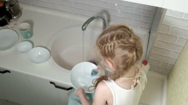 apronda güzel küçük kız bir sünger ile veliler yardım bulaşıkları parlak mutfakta bir sandalye üzerinde duruyor