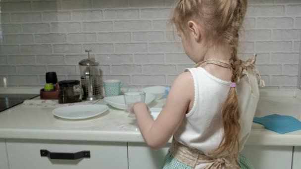 Belle petite fille dans un tablier est debout sur une chaise, dans la cuisine lumineuse, faire la vaisselle avec une éponge, aider les parents — Video