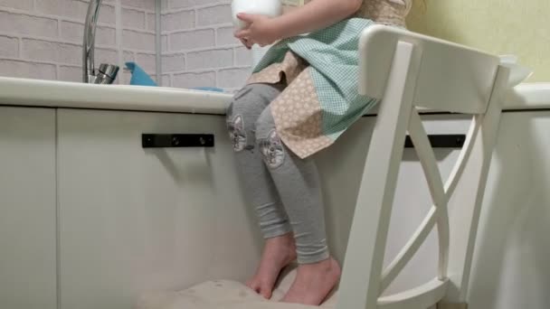 Mooi meisje in een schort is permanent op een stoel, in de lichte keuken, afwassen met een spons, ouders helpen — Stockvideo