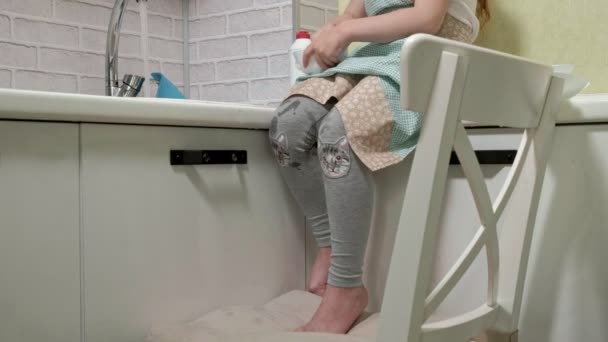 Vacker liten flicka i ett förkläde står på en stol, i det ljusa köket, gör rätter med en svamp, hjälper föräldrar — Stockvideo