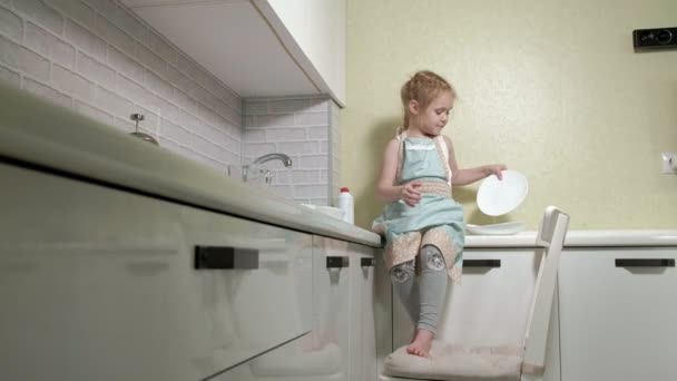 Beautiful little girl in an apron is standing on a chair in a bright kitchen, rinsing dishes with water, helping parents — Stock Video