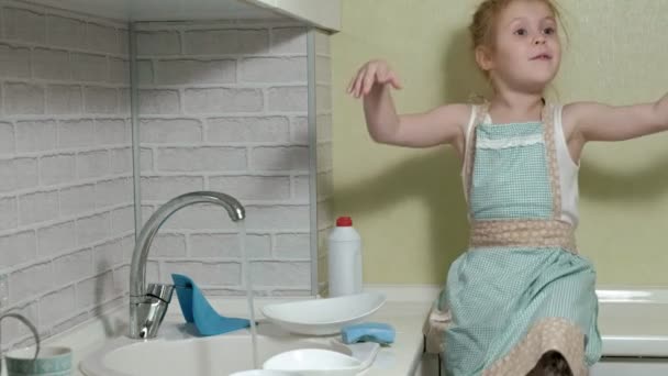 Belle petite fille dans un tablier est debout sur une chaise, dans la cuisine lumineuse, faire la vaisselle avec une éponge, aider les parents — Video