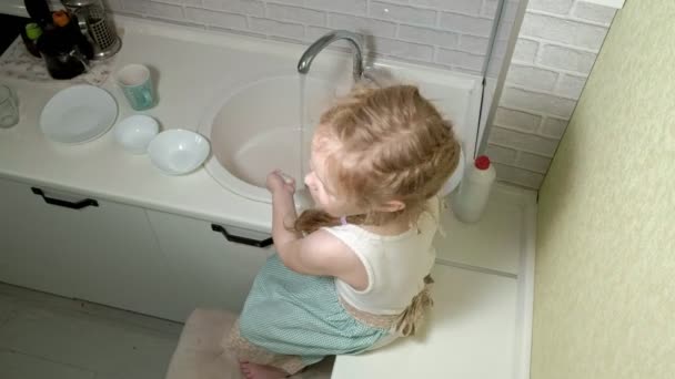 穿着围裙的漂亮小女孩站在椅子上, 在明亮的厨房里, 用海绵洗碗, 帮助父母 — 图库视频影像