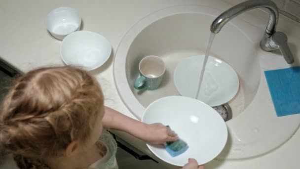 Mooi meisje in een schort is permanent op een stoel, in de lichte keuken, afwassen met een spons, ouders helpen — Stockvideo