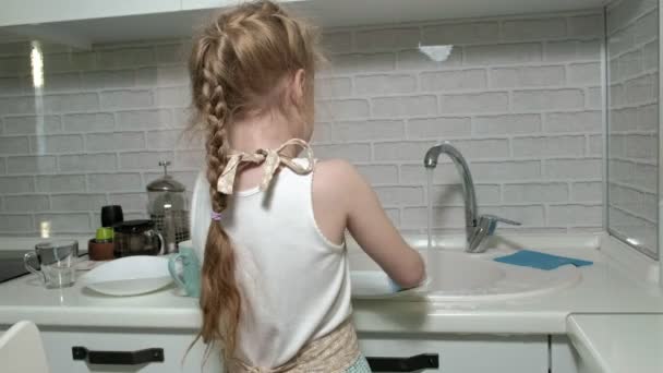 Όμορφο κοριτσάκι σε μια ποδιά στέκεται σε μια καρέκλα, στην φωτεινή κουζίνα, κάνει τα πιάτα με ένα σφουγγάρι, βοηθώντας τους γονείς — Αρχείο Βίντεο