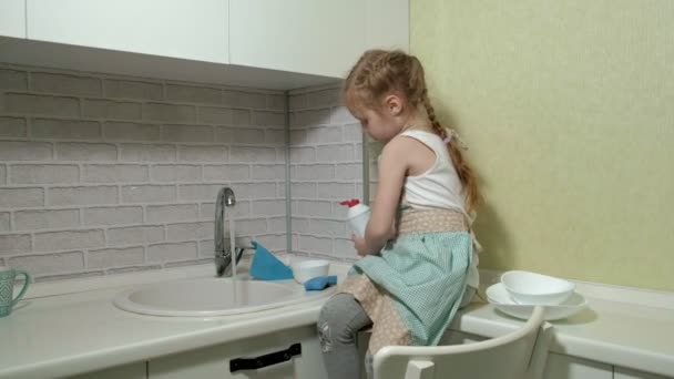 Красива маленька дівчинка в фартусі стоїть на стільці на яскравій кухні, застосовує миючий засіб для посуду до губки, допомагає батькам — стокове відео