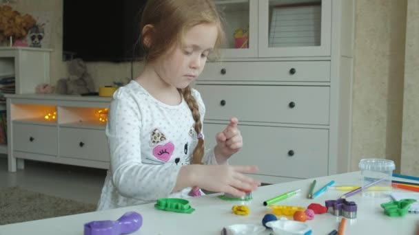 Маленькая девочка играет с пластилином, вырезает фигурки из формы, фигурки и цветные карандаши лежат на рабочем столе, развитие мелкой моторики рук — стоковое видео