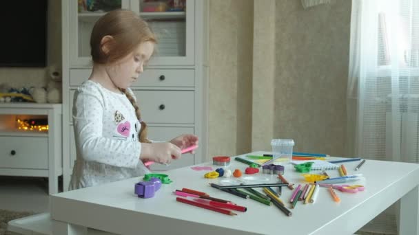 A little girl plays with plasticine, rolls it with her hands and cuts a figure, figures and colored pencils lie on the desktop, the development of fine motor skills of hands — Stock Video