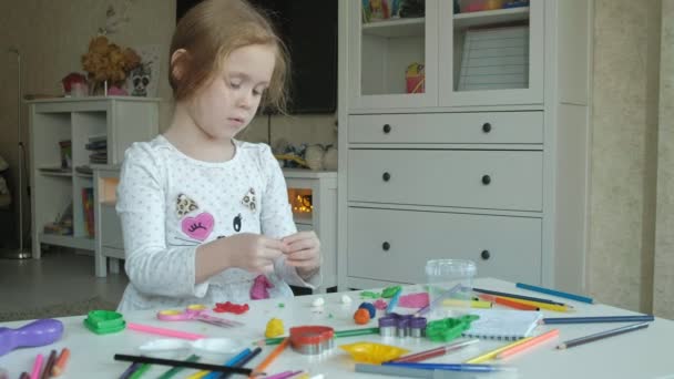 Счастливая маленькая девочка играет с пластилином, катит шарики руками, фигурки и цветные карандаши лежат на рабочем столе, развитие мелкой моторики рук — стоковое видео