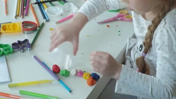Щаслива маленька дівчинка кладе різнокольоровий пластилін в ємність, на робочому столі фігури і кольорові олівці, розвиток тонких моторних навичок рук — стокове відео