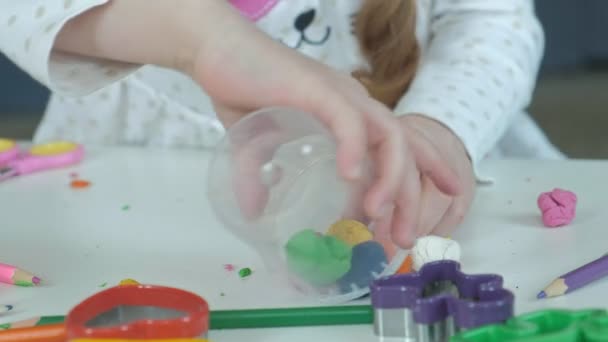 Щаслива маленька дівчинка кладе різнокольоровий пластилін в ємність, на робочому столі фігури і кольорові олівці, розвиток тонких моторних навичок рук — стокове відео