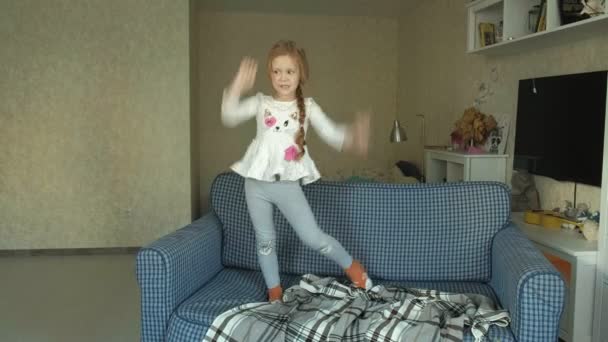 Маленькая девочка прыгает на диване в комнате, серая кошка садится рядом — стоковое видео