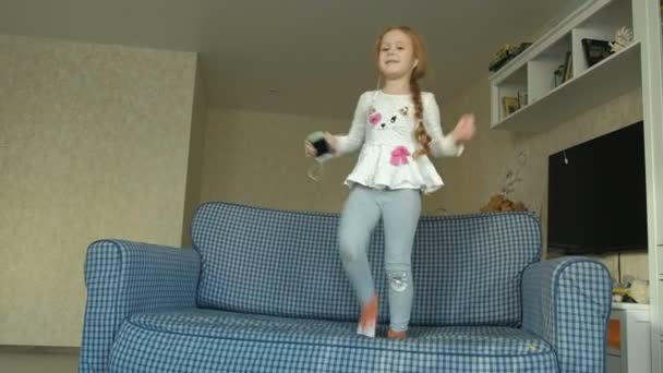 Meisje met een telefoon in haar handen en een koptelefoon springen, dansen op de sofa in de kamer — Stockvideo