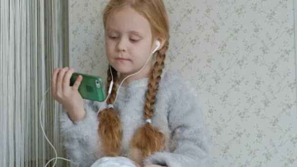 Glückliches kleines Mädchen mit Zöpfen im Kopfhörer schaut sich das Handyvideo an, sitzt in einem Raum auf der Fensterbank — Stockvideo
