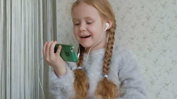 Happy little girl with pigtails in headphones looks at the phone video, sits in a room on the windowsill, hid a blanket — Stock Video